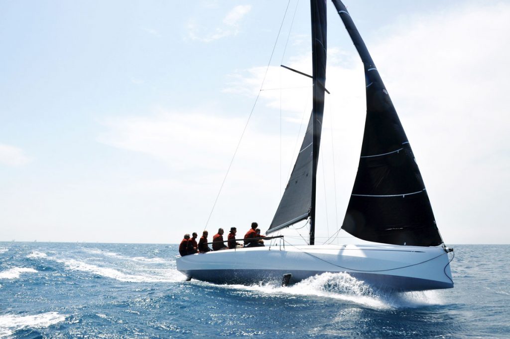 Crew sitting on the edge of a sail boat while it makes its way through the water.