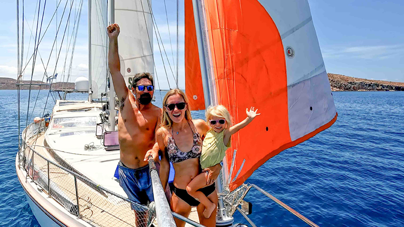 SV Delos crew smiling on deck while sailing in clear blue waters with bright orange sail raised.